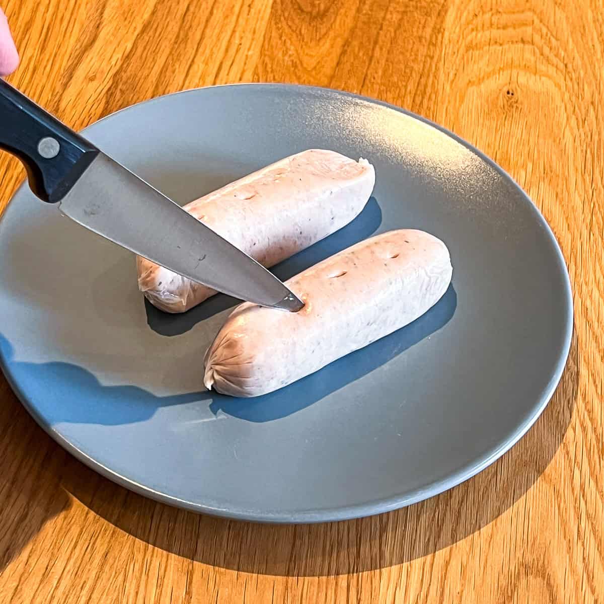 Two raw sausages on a grey plate being pricked with a knife.