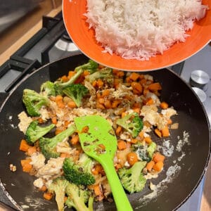 Rice being poured into a frying pan containing broccoli, carrots, onion and garlic.