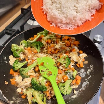 Rice being poured into a frying pan containing broccoli, carrots, onion and garlic.