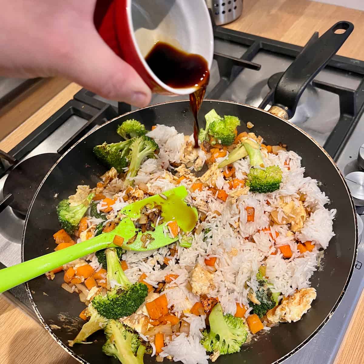 Pouring soy sauce into a frying pan of rice, broccoli, carrots, onion and garlic.
